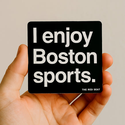 Hand holding a black square magnet with 'I enjoy Boston sports' text on a beige background