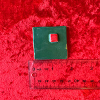 Hand holding a small green square handmade porcelain magnet with a red square and a ruler below for scale