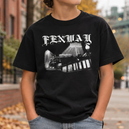 Kid wearing the Fenway Gothic youth tee on a city sidewalk in fall, ballpark light tower in the background