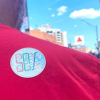 photo of a man with round sticker of design of the boston MBTA map in the shape of the word SOX, on his back with citgo sign in the distance.