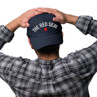 Person wearing a navy blue cap with 'The Red Seat' text, holding it behind their head on a white background.