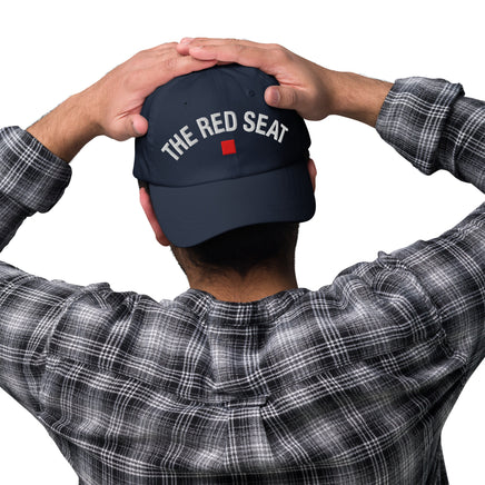 Person wearing a navy blue cap with 'The Red Seat' text, holding it behind their head on a white background.