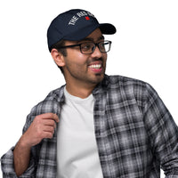 Man wearing a navy cap with 'The Red Bar' logo, plaid shirt, and glasses on a white background