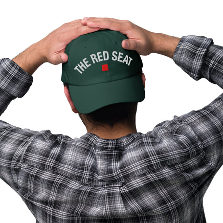 Person wearing a green cap with 'The Red Seat' text, holding it on a white background