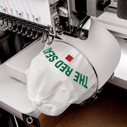 White cap with 'The Red Seat' text being embroidered on a machine.
