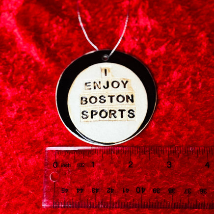 photo of round white and black porcelain ornament that says, i enjoy boston sports on red background with a ruler