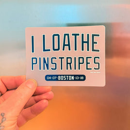 Refrigerator magnet with text 'I LOATHE PINSTRIPES' held by a person against a stainless steel fridge