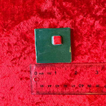 Hand holding a small green square handmade porcelain magnet with a red square and a ruler below for scale