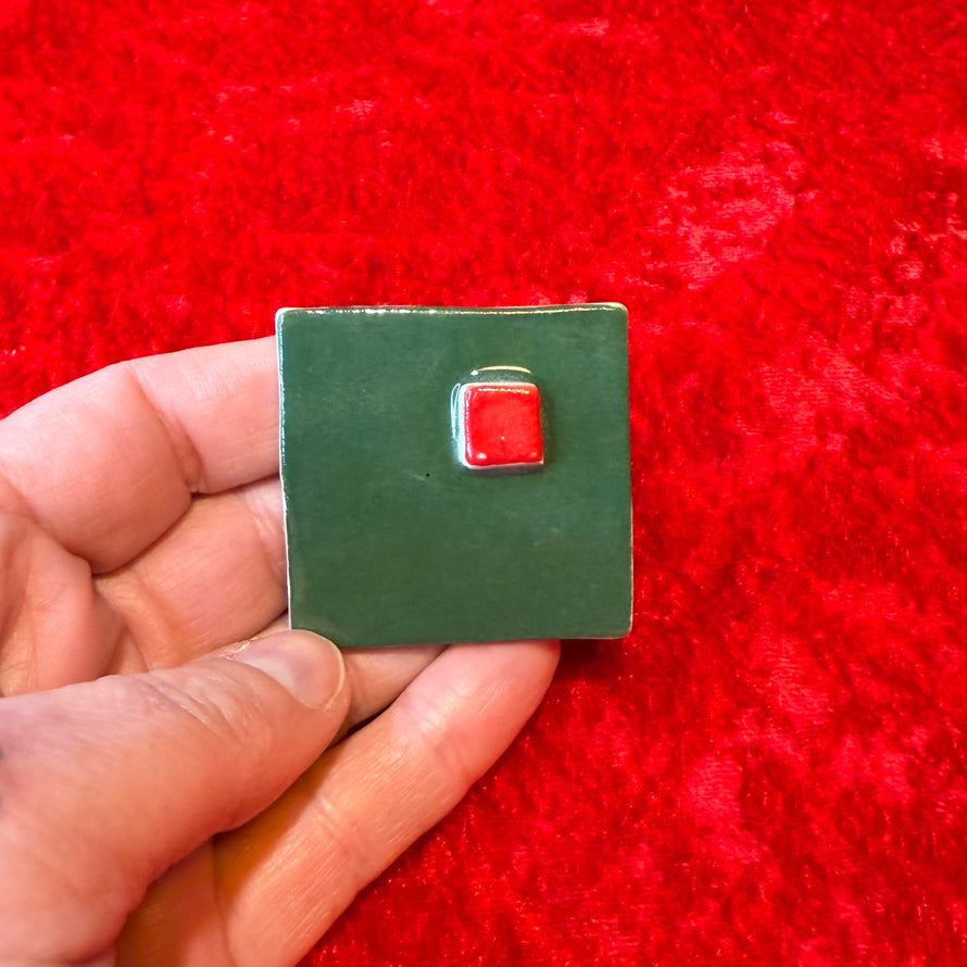 Hand holding a small green square handmade porcelain magnet with a red square